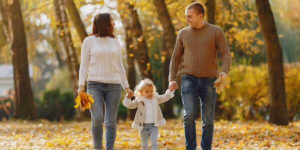 Two parents and a child walking on a path through a forest on a sunny fall day. The leaves on the trees have turned bright orange, yellwo and red.