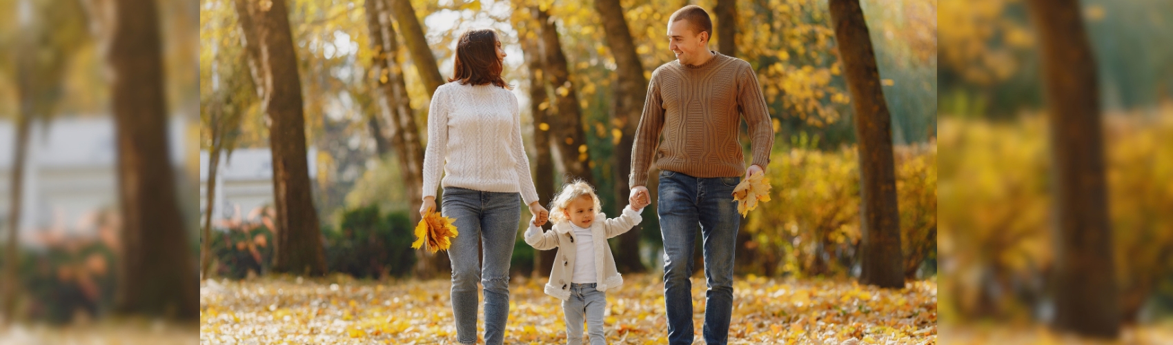 Two parents and a child walking on a path through a forest on a sunny fall day. The leaves on the trees have turned bright orange, yellwo and red.
