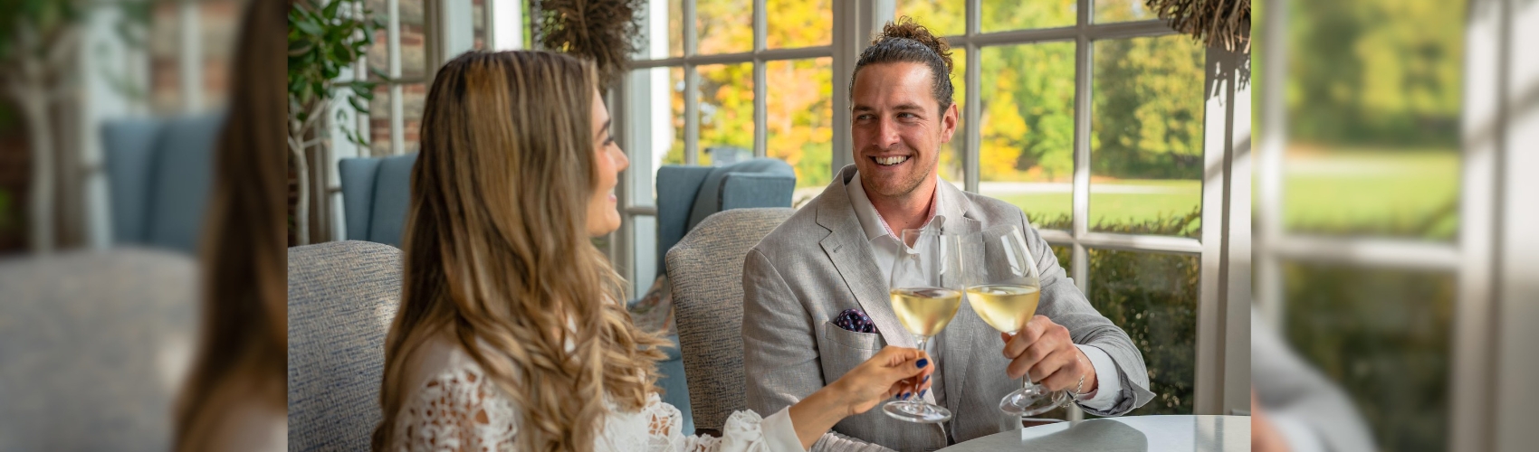A couple sitting at a window table in the restaurant at Langdon Hall in Cambridge. They are clinking their wine glasses. It is a sunny fall day outside